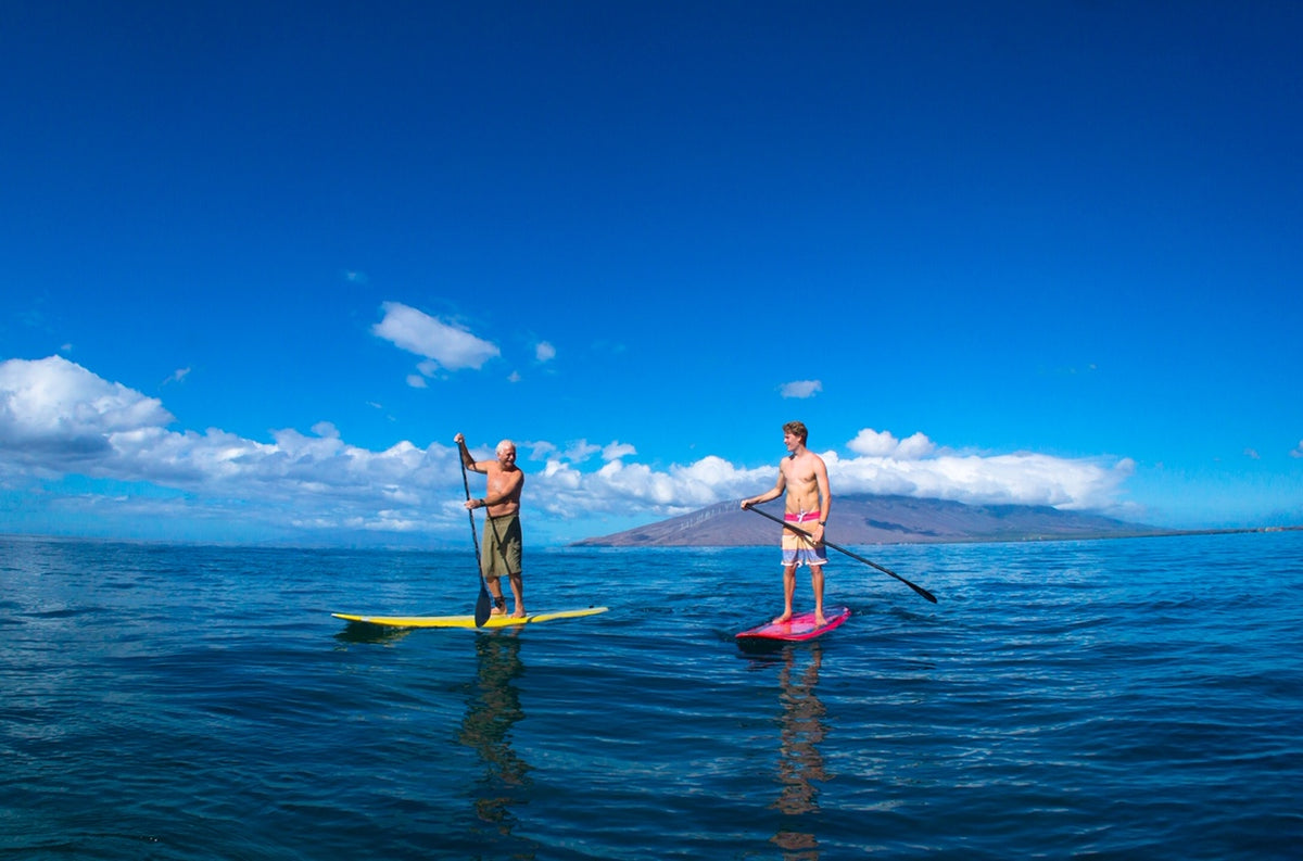 Two men stand on paddleboards on calm blue water under a bright blue sky with distant mountains and clouds. One board is yellow and the other is pink.