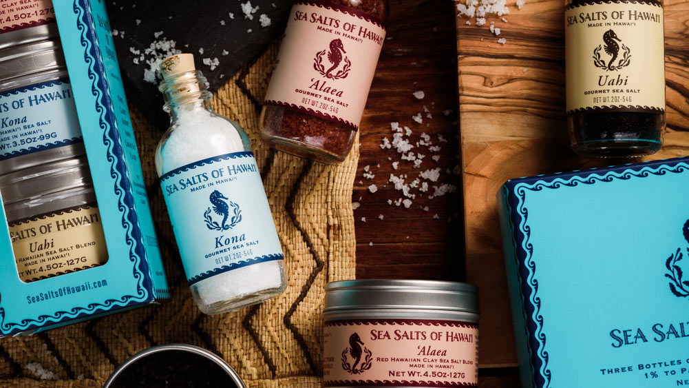 Sea Salt of Hawaii brand sea salt bottles and boxes arranged on a rustic wooden surface with coarse salt scattered around. Visible products include a boxed Uahi sea salt, a small bottle of Kona sea salt, a bottle of Alaea sea salt, and a tin of Uahi sea salt.
