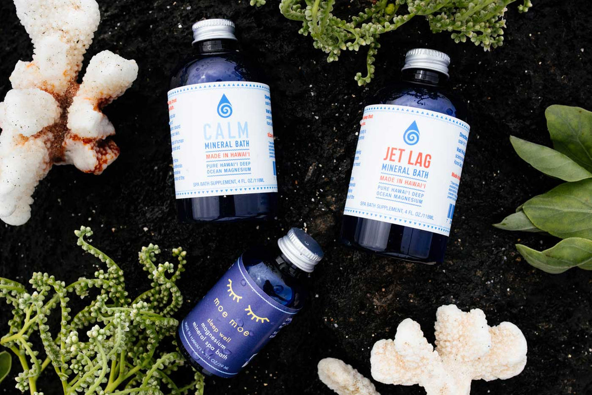 Three bottles of 'Calm' and 'Jet Lag' mineral baths on a dark surface with coral and plants.